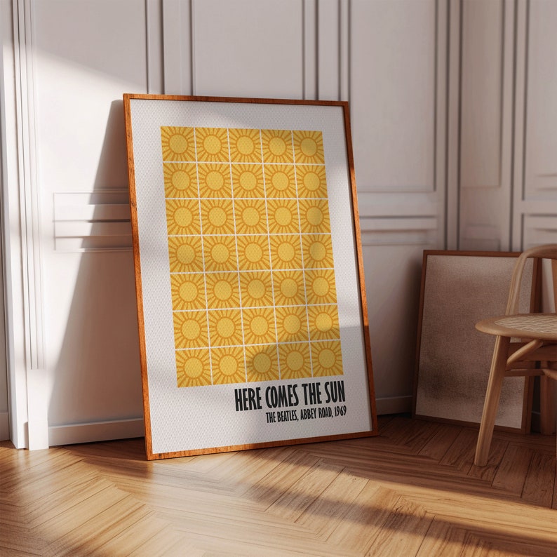 Peut inclure: Une affiche encadr&eacute;e avec un motif r&eacute;p&eacute;t&eacute; de soleils jaunes. Le texte "HERE COMES THE SUN" et "THE BEATLES, ABBEY ROAD, 1969" est affich&eacute; en noir. Le cadre est marron. L'affiche est pos&eacute;e contre un mur blanc, avec un sol en bois.
