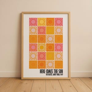 Peut inclure: Impression d'art encadr&eacute;e avec une grille d'illustrations de soleil dans des tons de rose, jaune et orange. Le texte "HERE COMES THE SUN" est affich&eacute; en dessous de la grille, avec "THE BEATLES, ABBEY ROAD, 1969" en dessous.
