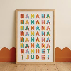 Può includere: Una stampa artistica incorniciata con le parole "NANANA NANANA NANANA NANANA NANANA NANHEY ! JUDE!" in lettere a blocchi 3D colorate. L'opera è in una cornice di legno chiaro, appoggiata a una parete color crema.