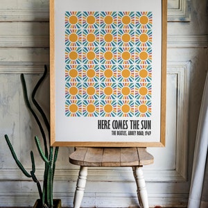 Peut inclure: Impression d'art encadr&eacute;e avec un motif r&eacute;p&eacute;t&eacute; de soleils jaunes et de rayons color&eacute;s. Le texte "HERE COMES THE SUN" et "THE BEATLES, ABBEY ROAD, 1969" sont imprim&eacute;s en dessous. L'impression est expos&eacute;e sur un tabouret en bois.