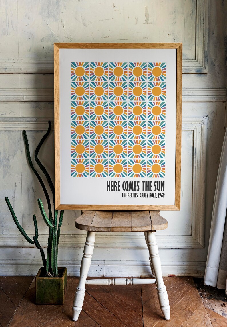 Peut inclure: Impression d'art encadr&eacute;e avec un motif r&eacute;p&eacute;t&eacute; de soleils jaunes et de rayons color&eacute;s. Le texte "HERE COMES THE SUN" et "THE BEATLES, ABBEY ROAD, 1969" sont imprim&eacute;s en dessous. L'impression est expos&eacute;e sur un tabouret en bois.