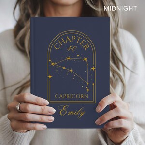 May include: A dark blue hardcover journal featuring a gold-colored arched frame containing a Capricorn constellation graphic, the text "CHAPTER 40", "CAPRICORN", and the name "Emily" in a cursive script. The word "MIDNIGHT" appears in the top corner.
