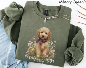May include: Military green sweatshirt with a graphic of a fluffy dog wearing a plaid scarf, surrounded by flowers. The text "Be Kind, Stay Fluffy" is printed below the dog. A silver necklace is draped on the sweatshirt.
