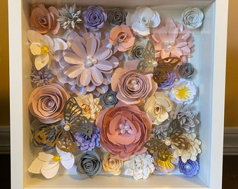 May include: A framed shadow box art piece featuring a variety of paper flowers in shades of pink, purple, gray, and white. The flowers are arranged in a cluster and include butterflies with pearl accents.