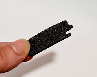 May include: A small, black, rectangular object with a textured surface and two notches on one end. The object is held between fingers against a white background.