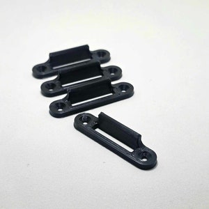 Może przedstawiać: Pięć czarnych plastikowych klipsów o prostokątnym kształcie z małą krawędzią po jednej stronie. Klipsy są zaprojektowane do trzymania przedmiotów w miejscu.