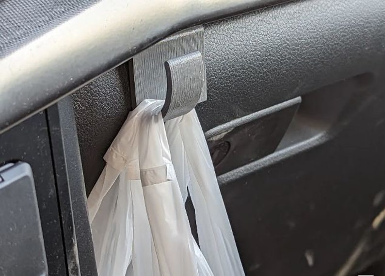 May include: A dark gray, 3D-printed hook attached to a car's dashboard. A white plastic bag hangs from the hook. The hook is designed to hold items in a vehicle.