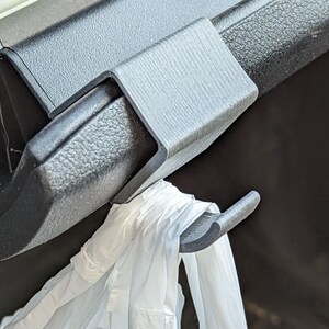 May include: A gray plastic car seat hook is attached to a black car interior component. A white plastic bag hangs from the hook. The hook is designed to hold items in a vehicle.