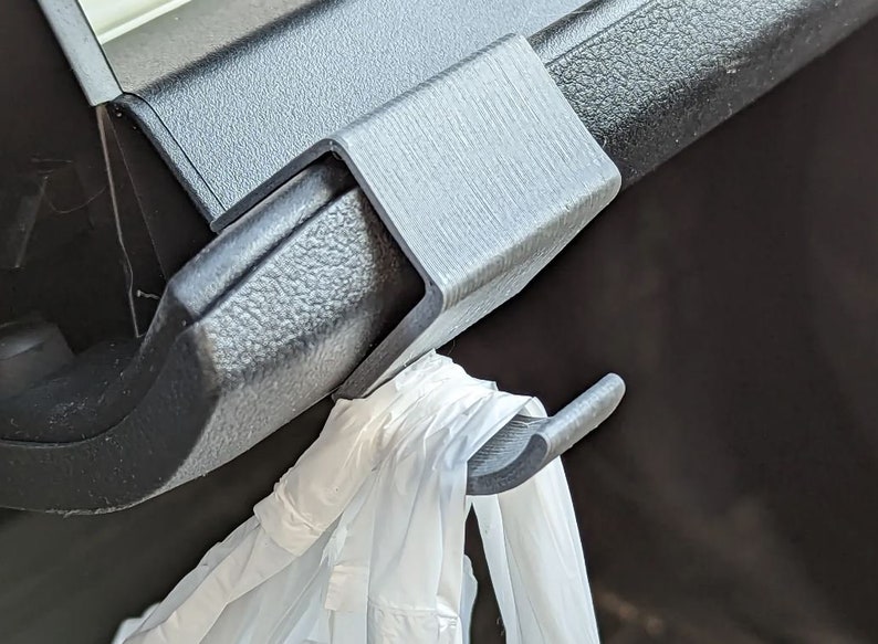May include: A gray plastic car seat hook is attached to a black car interior component. A white plastic bag hangs from the hook. The hook is designed to hold items in a vehicle.