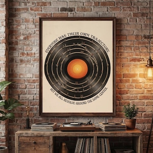 May include: A framed poster featuring a black vinyl record graphic with a bright orange centre. The text reads, "EVERYONE HAS THEIR OWN TRAJECTORY, BUT WE ALL REVOLVE AROUND THE SAME CENTRE." The design includes small figures walking along the record grooves.