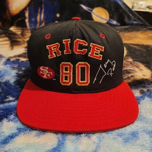 Puede incluir: Gorra de béisbol negra y roja con "RICE 80" bordado en rojo y dorado. La gorra presenta el logotipo de los 49ers de San Francisco y la firma de Jerry Rice. La visera es roja y la corona es negra.