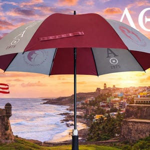 May include: A maroon, gray, and white umbrella with the Greek letters ΛΘΑ and the number 1975 printed on it. The umbrella is open, with a handle and a maroon strap. The background shows a coastal city and a sunset.
