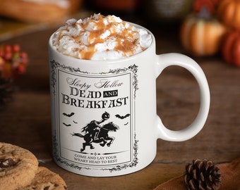 May include: White ceramic mug with a black and white illustration of a person riding a horse. The mug has the text "Sleepy Hollow Dead and Breakfast" and "Come and lay your weary head to rest". The mug is filled with a coffee drink topped with whipped cream and caramel.