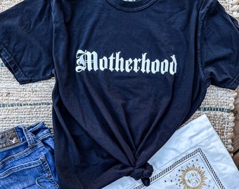 Könnte beinhalten: Ein schwarzes T-Shirt mit dem Wort "Mutterschaft" in weißen gotischen Buchstaben.
