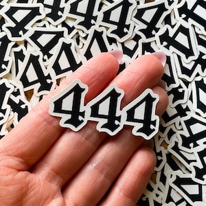 May include: A close-up of a hand holding three white stickers with the number "444" in a black gothic font. The stickers are surrounded by a pile of similar stickers, all with the same design.