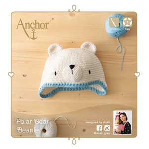 Op de afbeelding: Een handgemaakte, crèmekleurige gehaakte ijsbeermuts met een blauwe rand en zwarte gezichtskenmerken. De afbeelding bevat de tekst "Polar Bear Beanie" en "Anchor" met een haaknaald en eenvoudige instructies.