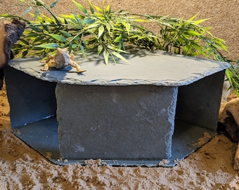 May include: A hexagonal slate hide with a small lizard on top. The hide is made of natural slate and has a rough texture. It is designed to provide a safe and secure hiding place for reptiles.