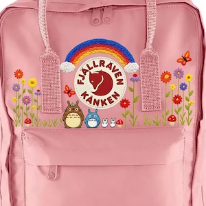 May include: A pink backpack with a top handle and front pocket. The backpack features an embroidered rainbow, flowers, butterflies, and cartoon characters. The brand logo is visible on the front.