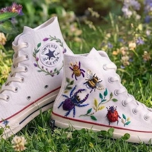 Könnte beinhalten: Ein Paar weiße High-Top-Sneaker aus Segeltuch mit gestickten Insektenmotiven, darunter eine Biene, ein Marienkäfer und ein Käfer auf einem Schuh sowie ein Blumenkranz um das Converse All Star-Logo auf dem anderen, mit rot-blauen Zierstreifen.