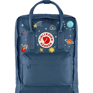 Puede incluir: Mochila azul marino con diseños bordados de temática espacial, incluyendo planetas, estrellas y un cohete. Cuenta con un bolsillo frontal, asa superior y el parche del logo "Fjallraven Kanken". La mochila mide aproximadamente 38 cm de alto.