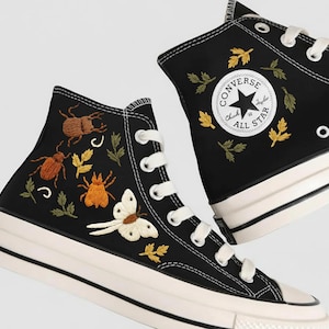 Könnte beinhalten: Schwarze High-Top-Sneaker mit gestickten Insekten- und Blattmotiven. Die Sneaker haben weiße Schnürsenkel, eine weiße Gummisohle und das Converse All Star-Logo. Die gestickten Details umfassen einen Schmetterling, Käfer und Blätter.