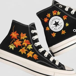 Op de afbeelding: Zwarte hoge sneakers met witte veters en zolen. De sneakers hebben geborduurde herfstbladeren in geel, oranje en rood. Eén schoen toont het Converse All Star-logo.