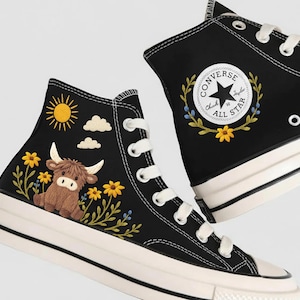 Puede incluir: Zapatillas altas negras con cordones y suelas blancas. Una zapatilla presenta una vaca de las Highlands marrón, flores amarillas, un sol y nubes. La otra tiene un logotipo de Converse All Star con detalles florales.
