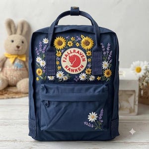 May include: A navy blue backpack featuring a circular Fjallraven Kanken logo patch on the front. The bag is decorated with embroidered yellow sunflowers, white daisies, and purple lavender sprigs surrounding the logo and on the front pocket.