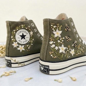 Peut inclure: Baskets montantes vert olive avec semelles et lacets blancs. Les baskets présentent des broderies florales et d'abeilles. Le logo Converse All Star est visible. Une option de chaussures unique et élégante.