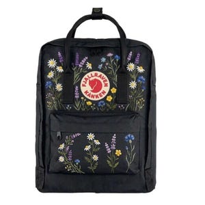 Könnte beinhalten: Schwarzer Rucksack mit gesticktem Blumendesign. Der Rucksack hat einen oberen Griff, eine Vordertasche und das Fjallraven-Logo. Die Blumenstickerei umfasst weiße Gänseblümchen, lila Lavendel und blaue Kornblumen.