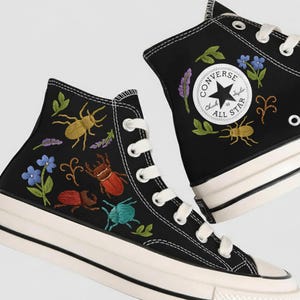 Könnte beinhalten: Schwarze High-Top Converse Chuck Taylor All Star Sneaker mit weißen Schnürsenkeln und Sohlen. Die Sneaker haben bunte, gestickte Insekten und florale Designs. Das Converse-Logo befindet sich an der Seite.