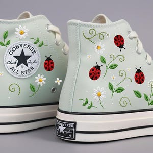 Könnte beinhalten: Paar mintgrüne High-Top-Sneaker mit Stickereien. Die Sneaker zeigen weiße Gänseblümchen, grüne Blätter und rote Marienkäfer. Das Converse All Star-Logo ist sichtbar.
