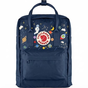 Puede incluir: Una mochila azul marino con un parche circular del logotipo de Fjällräven Kånken en el centro. El exterior está decorado con un estampado de temática espacial que incluye un astronauta, un cohete, planetas, estrellas y constelaciones.