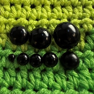 Puede incluir: Una colecci&oacute;n de ojos de seguridad negros brillantes de varios tama&ntilde;os, dispuestos sobre un fondo de ganchillo verde y amarillo vibrante. Los ojos var&iacute;an en tama&ntilde;o, adecuados para varios proyectos hechos a mano.