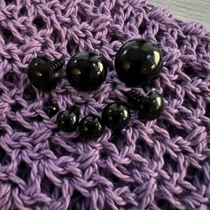 Puede incluir: Una colecci&oacute;n de ojos de seguridad negros de varios tama&ntilde;os, algunos con postes atornillables, dispuestos sobre una superficie de ganchillo lavanda texturizada. Los ojos brillantes son redondos y lisos, perfectos para proyectos de manualidades.