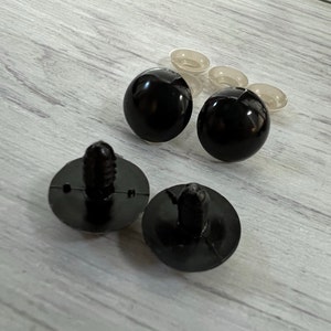 Puede incluir: Una colecci&oacute;n de suministros de artesan&iacute;a de pl&aacute;stico negro, que incluye dos esferas negras redondas y brillantes, dos sujetadores negros con una base circular y varias piezas peque&ntilde;as de pl&aacute;stico transparente. Los art&iacute;culos est&aacute;n sobre una superficie de madera clara.