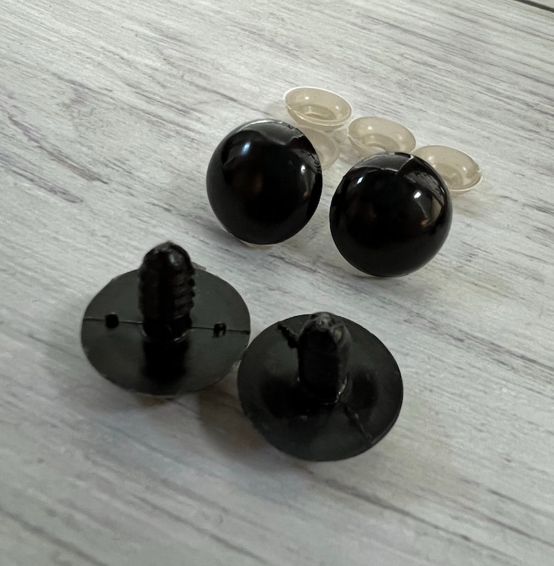 Puede incluir: Una colecci&oacute;n de suministros de artesan&iacute;a de pl&aacute;stico negro, que incluye dos esferas negras redondas y brillantes, dos sujetadores negros con una base circular y varias piezas peque&ntilde;as de pl&aacute;stico transparente. Los art&iacute;culos est&aacute;n sobre una superficie de madera clara.