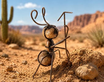 May include: A metal sculpture of an ant with a rock body and wire legs and antennae. The ant is standing on a small pile of dirt in a desert landscape with a cactus and mountains in the background. The sculpture is a unique piece of art.