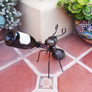 May include: A whimsical metal ant wine bottle holder. The ant-shaped sculpture is made of dark metal and holds a wine bottle. The ant has a body made of rounded stones and wire legs and antennae. The bottle is dark with a white label.