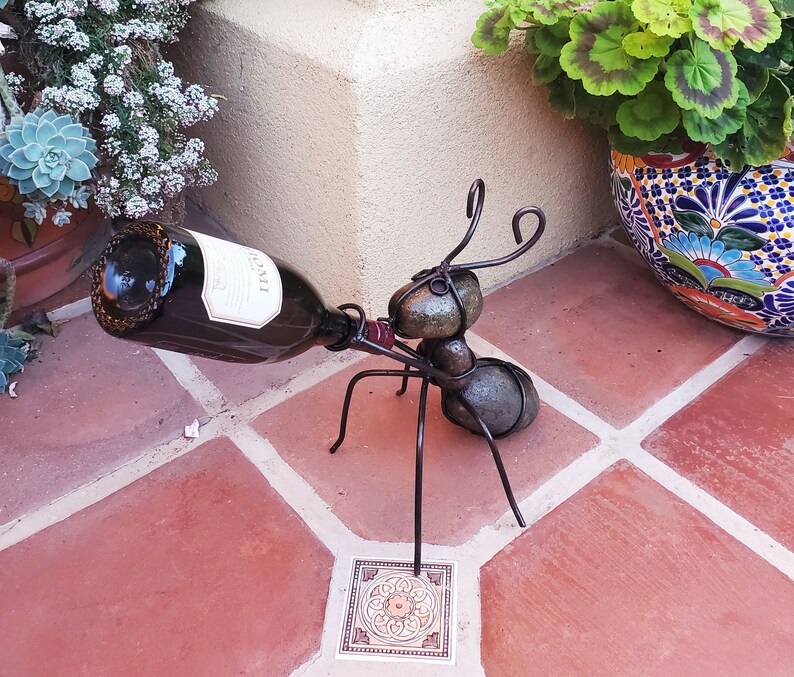 May include: A whimsical metal ant wine bottle holder. The ant-shaped sculpture is made of dark metal and holds a wine bottle. The ant has a body made of rounded stones and wire legs and antennae. The bottle is dark with a white label.
