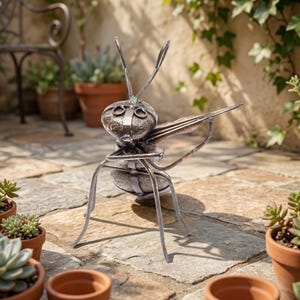 May include: A metal sculpture of an insect with a detailed body, wings, and antennae. The sculpture is made of silver-colored metal and is positioned on a stone patio, surrounded by potted plants. The insect sculpture is a decorative garden accent.