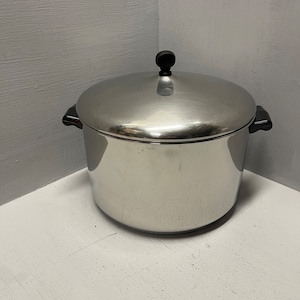 May include: A large, stainless steel stock pot with a black lid and handles. The pot is sitting on a white surface.