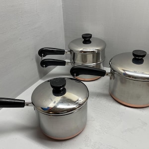 May include: A set of three stainless steel saucepans with copper-colored bases and black handles. Each pot features a matching stainless steel lid with a black knob. The pots vary in size and are arranged together, showcasing a polished metallic finish.