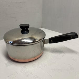 May include: A stainless steel saucepan with a copper-colored base and a black handle. The pot has a lid with a black knob. The pan is designed for cooking and is suitable for various kitchen tasks.