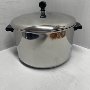 May include: A stainless steel cooking pot with a lid. The pot has two black handles and a black knob on the lid. The pot is sitting on a white surface.
