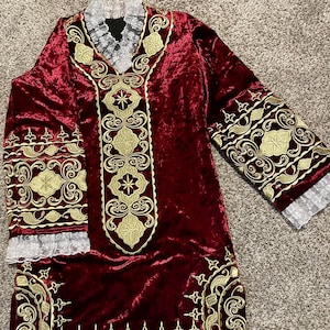 May include: A long, burgundy velvet robe with gold embroidery. The robe has a V-neckline and long sleeves with lace trim. The embroidery features intricate floral and geometric patterns.