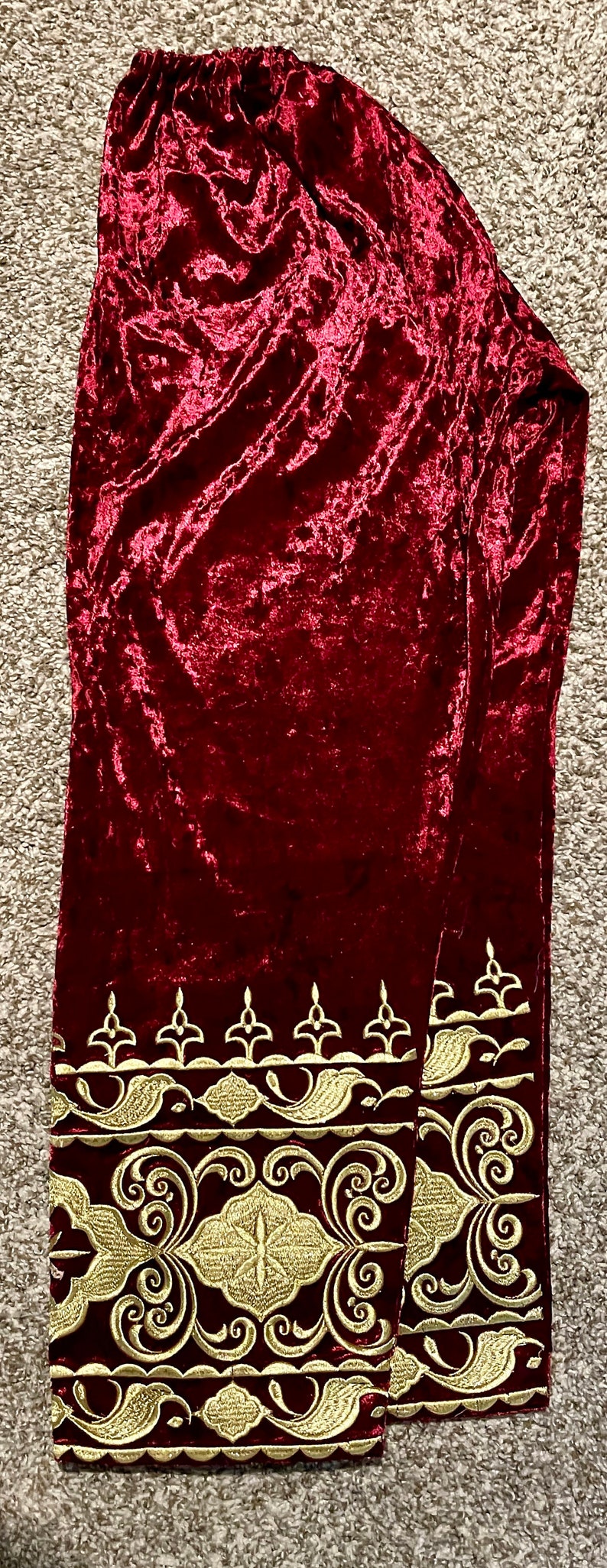 May include: A pair of burgundy velvet pants with gold embroidered trim. The trim features a repeating pattern of stylized flowers and leaves.