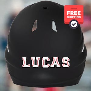 May include: A black baseball helmet with the name "LUCAS" in white letters with red baseball stitching. The helmet has ventilation holes and a sticker that says "FREE SHIPPING".