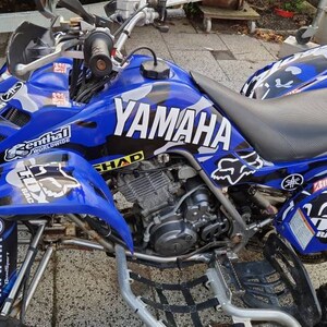 May include: A blue Yamaha ATV with a black and white camouflage design. The ATV has a black seat, black handlebars, and black wheels. The ATV has a Yamaha logo on the side and a Fox Racing logo on the back.