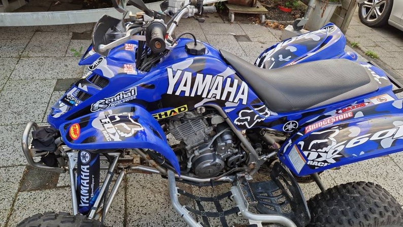 May include: A blue Yamaha ATV with a black and white camouflage design. The ATV has a black seat, black handlebars, and black wheels. The ATV has a Yamaha logo on the side and a Fox Racing logo on the back.